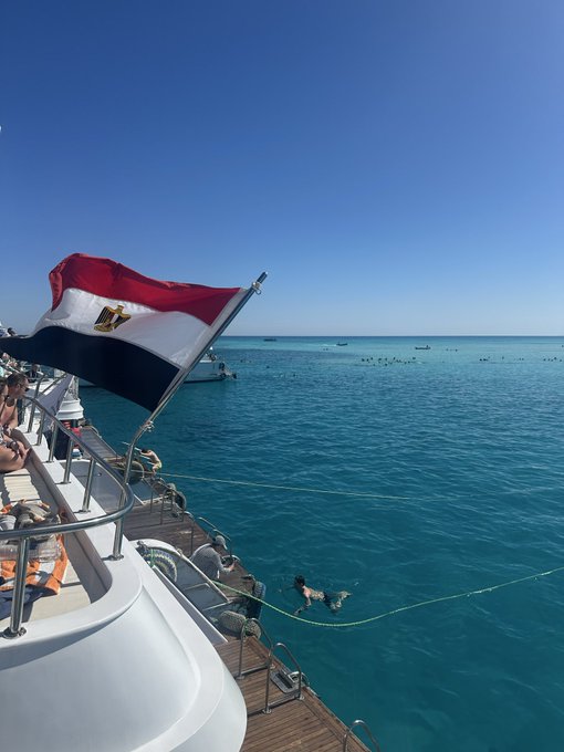 An Egyptian flag waves from a boat on turquoise waters as swimmers enjoy the Red Sea, capturing a perfect "Good Morning" scene from a sunny seaside getaway.