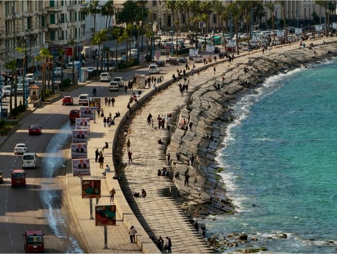 An elevated view of a coastal promenade lined with palm trees, billboards, vehicles, and pedestrians walking alongside a stone seawall next to turquoise ocean waters.