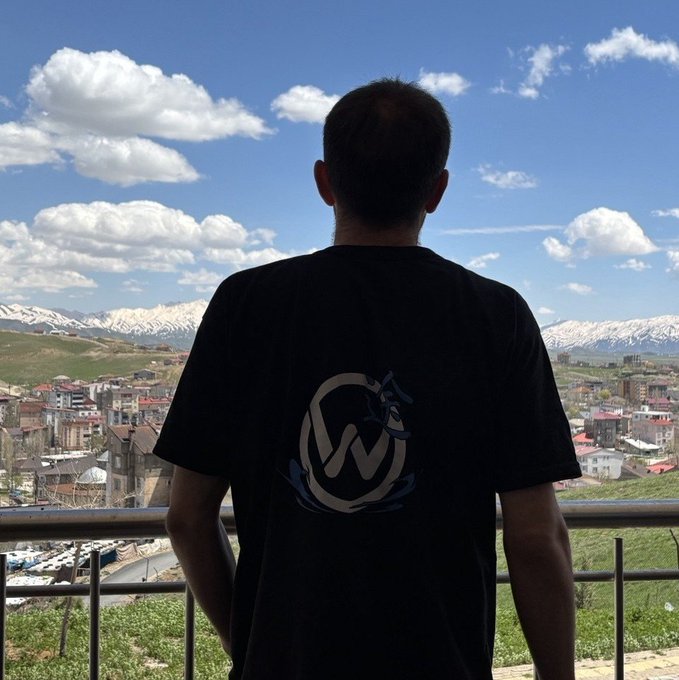 A man in a black t-shirt with a stylized 'W' wolf logo stands looking out over a town nestled below snow-capped mountains under a partly cloudy sky.
