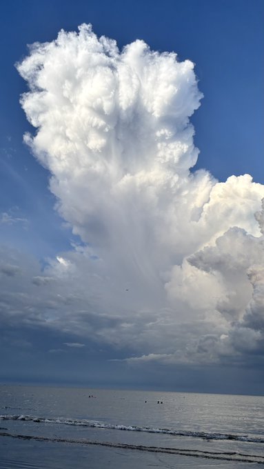 A dramatic cumulus cloud formation towers over the ocean, casting streaks of rain toward the water below.