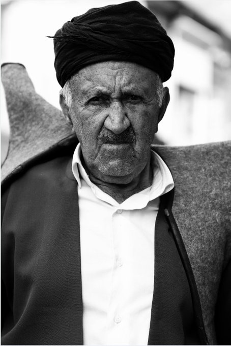 A black-and-white portrait of an elderly man wearing a dark turban and a white shirt under a dark jacket, showing a weathered face with a serious expression.