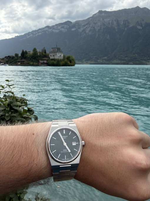 A man holds up his wrist to showcase his new Tissot PRX watch against a scenic alpine lake and mountain backdrop, celebrating the timepiece while referencing $NBIS as a potential top stock pick. The watch's texturized blue dial and integrated bracelet stand out prominently in the foreground, complementing the cool tones of the surrounding water and distant peaks.