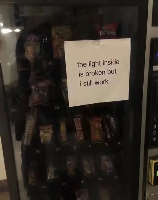 A vending machine with a handwritten sign reading 'the light inside is broken but i still work' displays various snacks including Doritos, Cheetos, and KitKat.