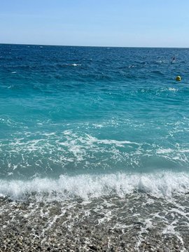 A coastal scene with turquoise water and white waves crashing onto a pebble beach, with people swimming in the distance under a clear blue sky.