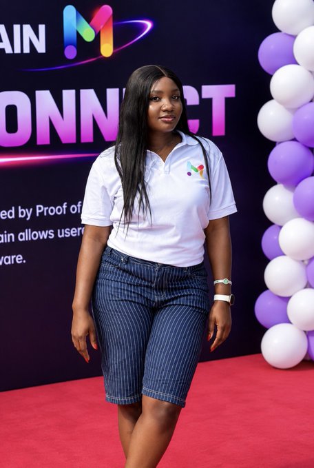 A woman stands on a red carpet wearing a white polo shirt and blue striped bermuda shorts, posing in front of a backdrop with partial text 'CONNECT' and 'ed by Proof of' alongside purple and white balloons.