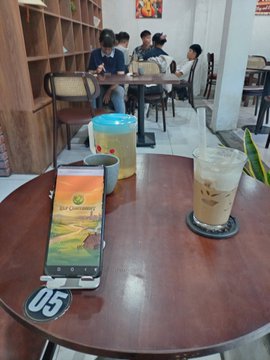 The image shows a wooden table with various items placed on top of it, including a cell phone displaying a game logo. The table also has two cups - one filled with coffee and another containing an unknown beverage. There are three people sitting at separate tables in the background, engaged in their own activities. A chair is visible near the left side of the image.