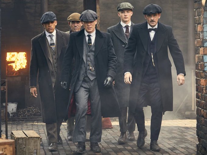 Five men in period clothing—flat caps, overcoats, three-piece suits, and ties—walk purposefully down a cobblestone alleyway with fire visible in the background, evoking the Peaky Blinders aesthetic.
