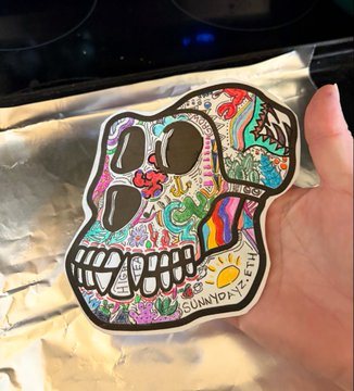 A hand holds up a colorful sticker featuring a decorated sugar skull design with intricate patterns and the word 'SUMMER' at the bottom.