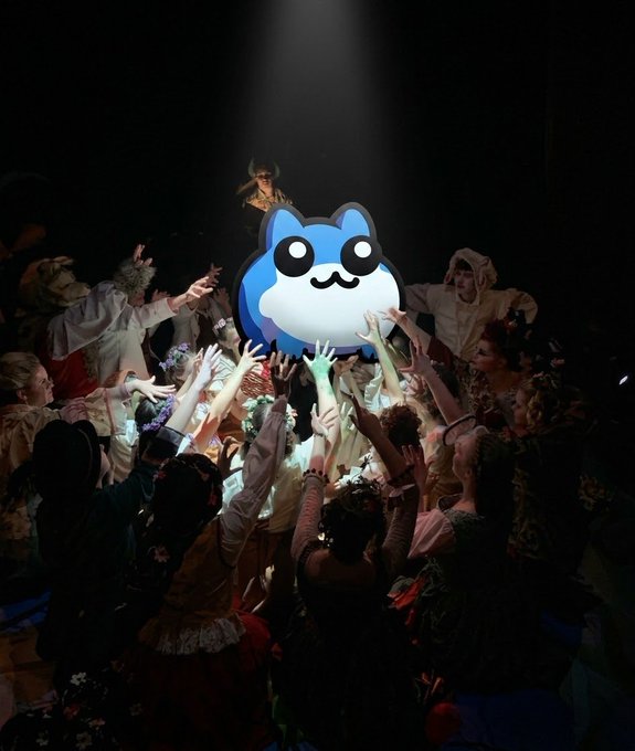 A group of performers in elaborate costumes reach up toward a large illuminated blue cat mascot head on a stage with a spotlight overhead.