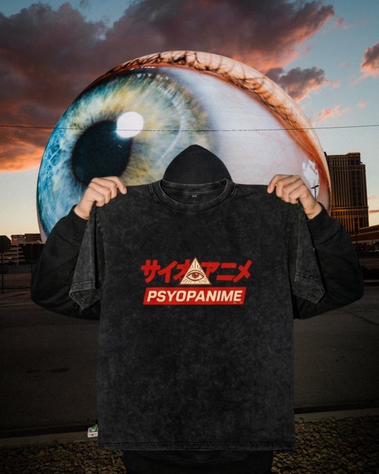 A person wearing a black hoodie holds up a distressed black t-shirt displaying "サイコアニメ PSYOPANIME" in red text, with a large, surreal eyeball visible in the sky behind them.