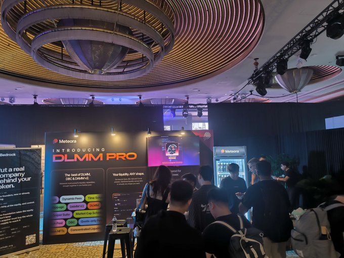 An indoor event or conference with attendees gathered around a booth promoting "DLMM PRO" by Meteora, featuring a large decorative spiral light fixture on the ceiling.