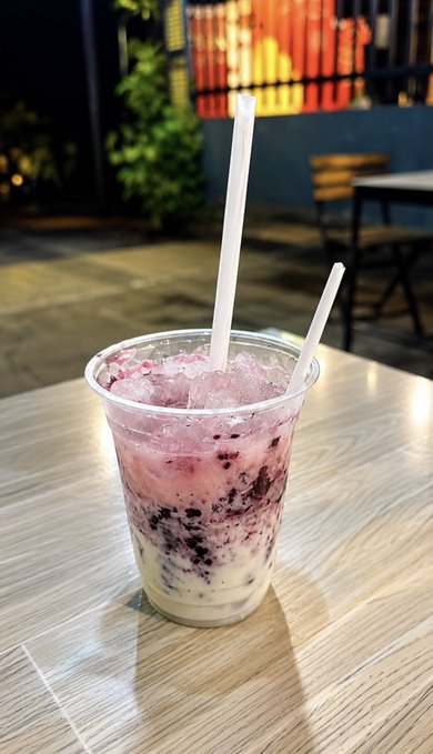 A plastic cup filled with a layered iced beverage featuring purple/blue berries on top and a creamy white layer below, served with two white straws on a wooden table.