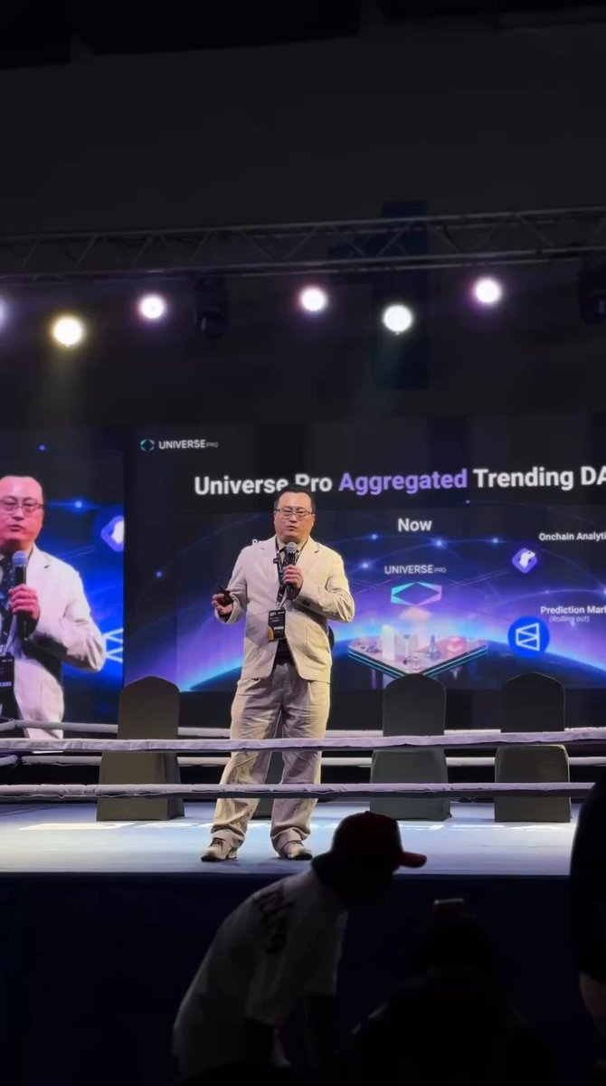 The image features a man standing at a podium, giving a speech or presentation. The background of the scene includes a large screen displaying information about the Universe Pro ecosystem. There are also several chairs placed around the stage for attendees to sit and listen to the speaker.