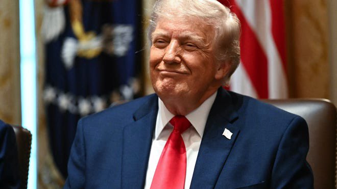 The image features a man wearing a suit, tie, and red shirt sitting at a table with his hands together. He has a smile on his face as he looks directly at the camera. The background of the image includes an American flag and a blue curtain, suggesting that this scene takes place in an official setting or within a government building. There is also text present in the image that reads "I told you guys Lock in. $XRP". This message seems to be related to the man's suit and tie, possibly indicating his position as a high-ranking official or someone of significant importance.