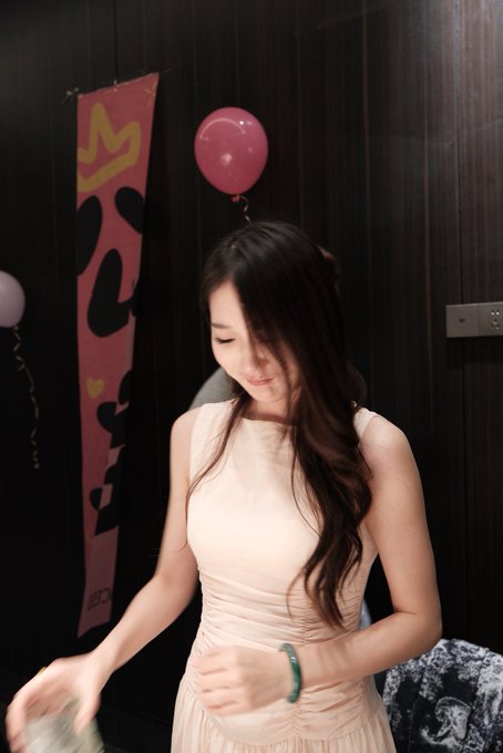 A young woman with long dark hair looks down while wearing a light pink sleeveless dress and a green jade bracelet, standing near a dark wood-paneled wall decorated with a pink banner and balloons.