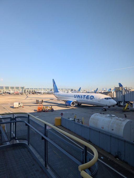 A United Airlines aircraft sits at the gate, with "UNITED" prominently displayed on the fuselage, as a traveler prepares to head to BitcoinConf Asia. The scene captures the journey's start at the airport before heading to the conference.