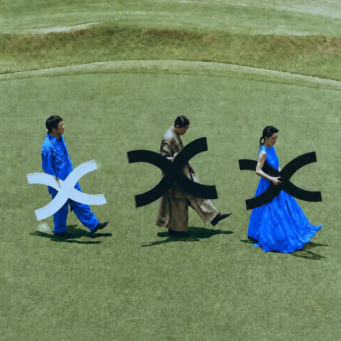 Three people walk across a grassy field carrying large cross-shaped symbols—one white and two black—symbolizing the cultural burden referenced in the post, where each figure shoulders a piece of the weight through their bold attire and solemn stride.