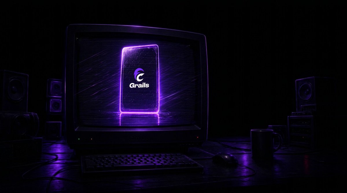 The image shows a computer monitor with a purple logo displayed on its screen. The monitor is placed on a desk next to a keyboard and mouse. In front of the monitor, there are speakers that appear to be turned off or muted. A cup can also be seen in the scene. The background features a dark room with a television set located behind the computer setup.