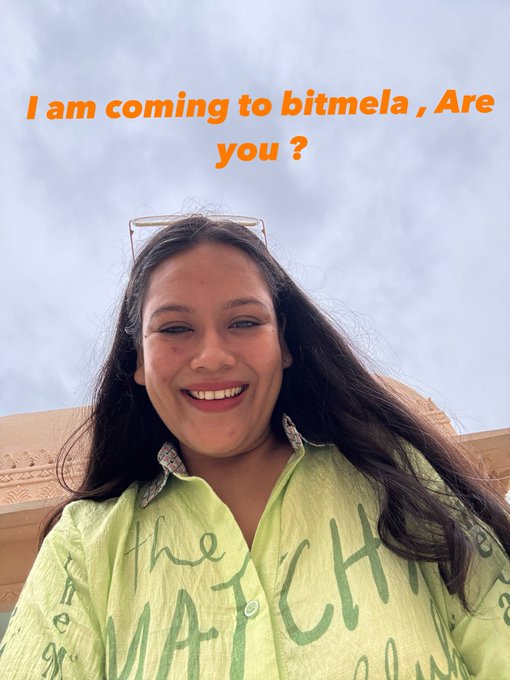 A selfie-style photo of a smiling young woman wearing a green shirt with text and sunglasses on her head, with orange overlaid text reading 'I am coming to bitmela , Are you ?'