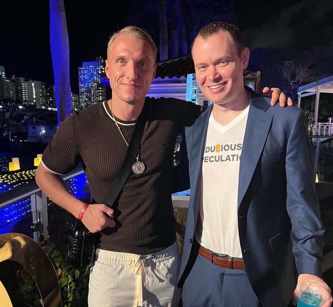 Two smiling men pose together at an outdoor evening event with city lights in the background; one wears a navy blazer over a white t-shirt reading "DUBIOUS CALCULATI..." while the other sports a ribbed brown shirt and a camera strap, playfully embodying the caption's "Haters gonna hate. Potatoes gonna Potate." sentiment with their cheerful, unbothered camaraderie.
