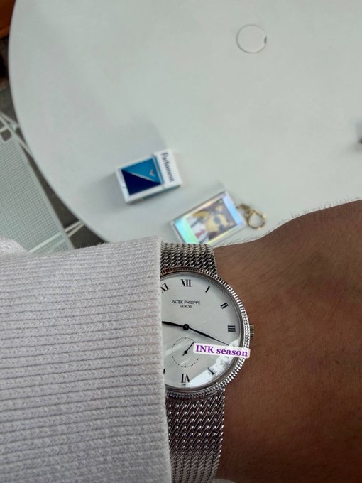 A person's wrist wearing a Patek Philippe Geneve watch with Roman numerals and a mesh band, positioned near a white table with a small blue and white object and a keychain, with 'INK season' text visible on the watch.