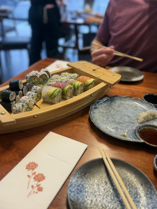 A wooden boat-shaped platter filled with assorted sushi rolls sits on a dining table alongside ceramic plates and chopsticks.