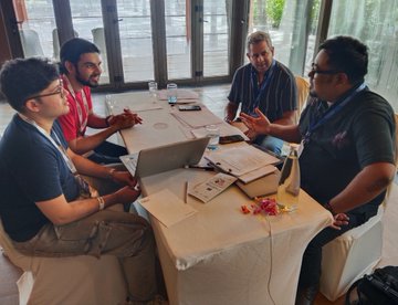 The image shows a group of four men sitting around a table with laptops open, engaged in a discussion or working together on a project. The room has large windows that allow natural light into the space, creating an atmosphere conducive for collaboration and creativity. The men are focused on their work, suggesting they have been working diligently to come up with innovative solutions. The post about the AlgoBharat Hack Series highlights the importance of teamwork and problem-solving in today's competitive business environment.