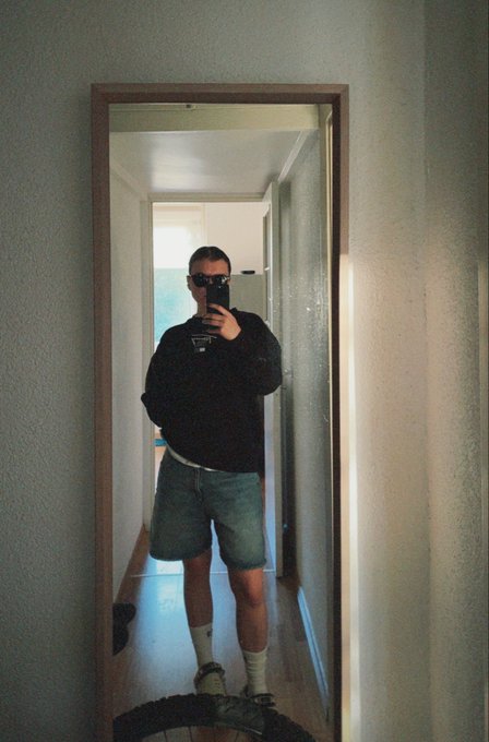 A man wearing sunglasses, a dark jacket, and denim shorts takes a mirror selfie in a hallway while holding a camera.
