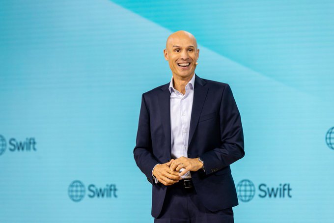 A bald man in a navy suit smiles while standing on stage in front of a light blue backdrop displaying the "Swift" logo, appearing to give a presentation. The image relates to the post announcing SWIFT's official initiative to connect its network of over 11,000 banks to the crypto world.