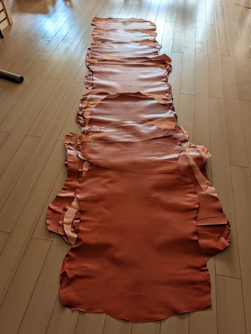 Several large pieces of tan and brown leather hide are laid out in a row on a light wooden floor, appearing to be prepared for cutting.