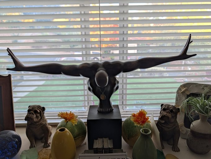 A windowsill display featuring a bronze bust sculpture of a horned, headless figure with outstretched arms, flanked by two bulldog figurines, vases with flowers, and a view of a yard through the blinds.