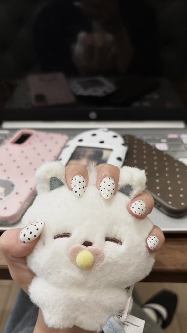 A hand with white and black dalmatian-spotted nail art holding a small plush white bear toy, with a laptop and pink phone case in the background.