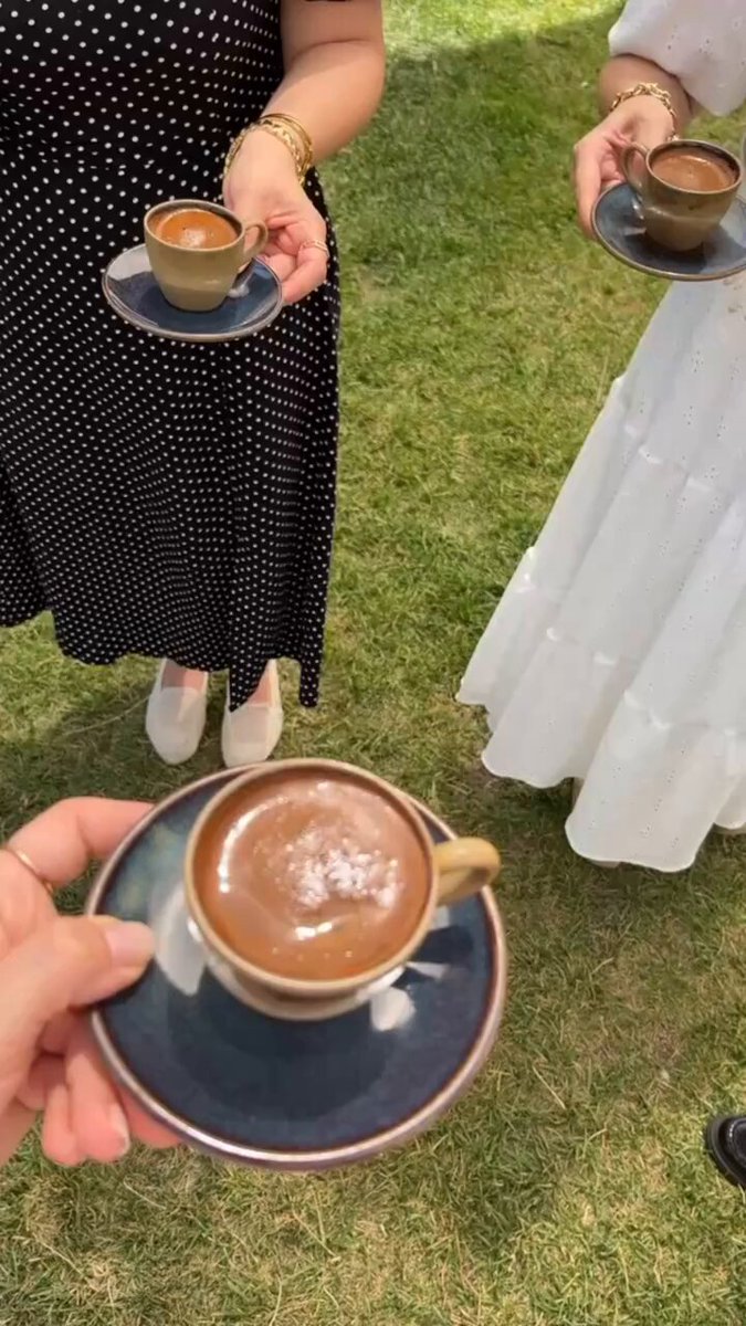 The image shows three people standing together, each holding a cup of coffee. The cups are placed on saucers or plates, which are blue in color. One person has their hand raised towards another person's cup, possibly offering a taste test or sharing a moment with friends. The background features grass and trees, creating an outdoor setting for the gathering.