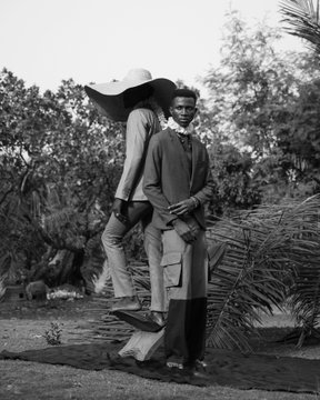 A black-and-white photograph of two people outdoors, one wearing a wide-brimmed hat standing behind a seated young man in a robe, surrounded by tropical foliage.