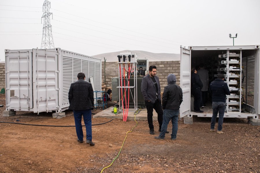 Several workers inspect electrical equipment in white shipping containers at an outdoor industrial site with a power transmission tower visible in the background. The image does not depict crypto mining or financial activities but rather shows physical infrastructure—likely a substation or generator setup—that may relate to the energy-intensive nature of cryptocurrency mining operations referenced in the post about Iran using crypto to bypass U.S. sanctions.