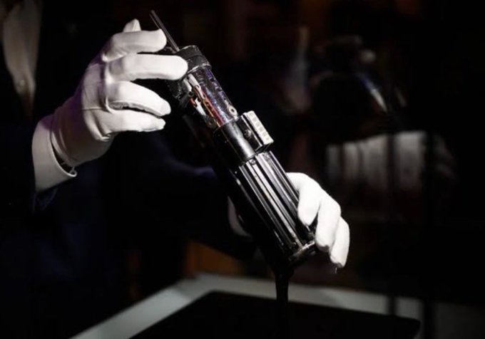 A person wearing white gloves carefully pours a thick black liquid from a metallic cylindrical device in a dimly lit setting.