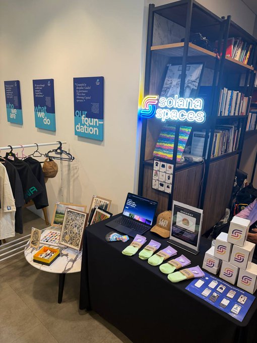 A photo of a Solana Spaces merchandise booth displaying t-shirts on hangers, stickers, socks, hats, and a laptop on a table with a neon 'solana spaces' sign on a bookshelf, alongside posters reading 'who we are', 'what we do', and 'our foundation'.