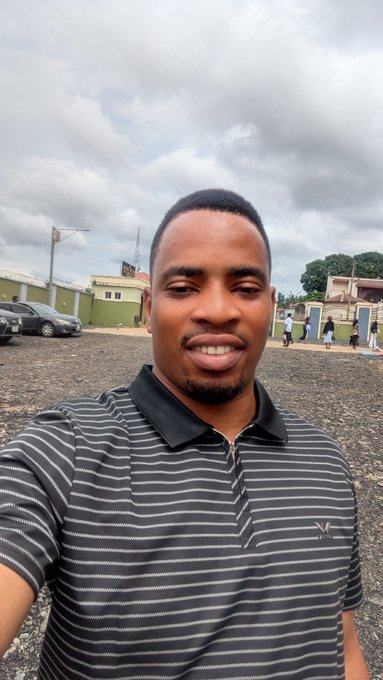 A selfie of a smiling young man wearing a striped black polo shirt, taken outdoors with a cloudy sky, parked cars, and buildings visible in the background.