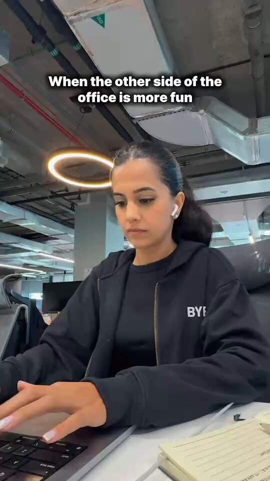The image features a woman sitting at a desk, typing on a laptop computer while wearing a black sweatshirt with white text that says "BYBIT". The background of the image shows an office setting with a ceiling fan visible above her head. The caption of the image reads "When the other side of the office is more fun #Bybit #NewFinancialPlatform", which seems to be related to the content displayed on the laptop screen, possibly discussing financial matters or trends in the industry.