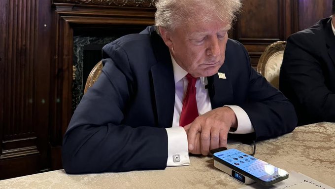 Donald Trump looks down at a smartphone displaying what appears to be stock market data while seated at an ornate table, visually reinforcing the post's claim about his extensive 1,156 stock trades worth up to $270 million in July—highlighting the irony of a stock-trading ban that covers Congress but excludes the President.