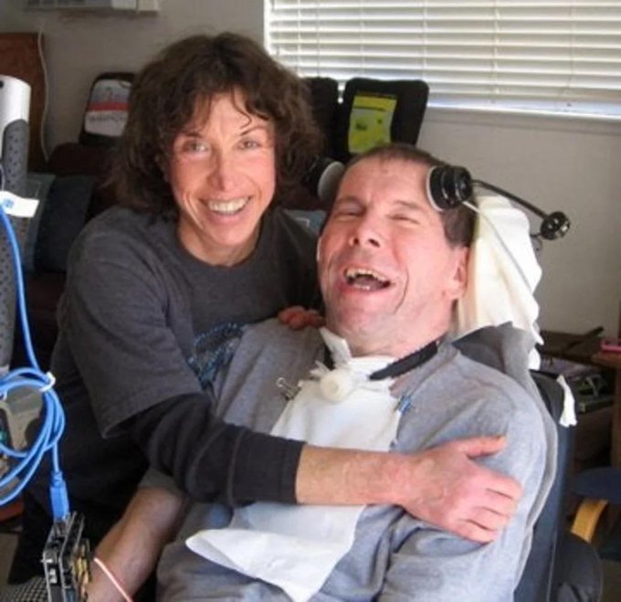 A heartwarming photo of a smiling woman hugging a man who appears to have disabilities, seated in what looks like a medical or care setting.