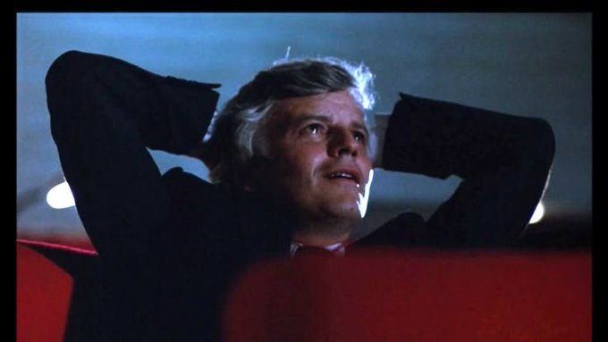 A still from a film showing a man with gray hair leaning back in red theater seats, hands behind his head.