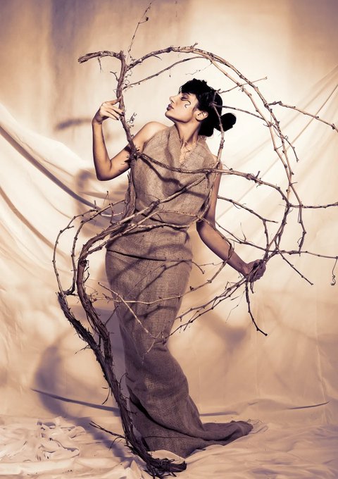 A woman in a flowing, neutral-toned gown poses elegantly with intertwined branches in a sepia-toned, artistic portrait against draped fabric.