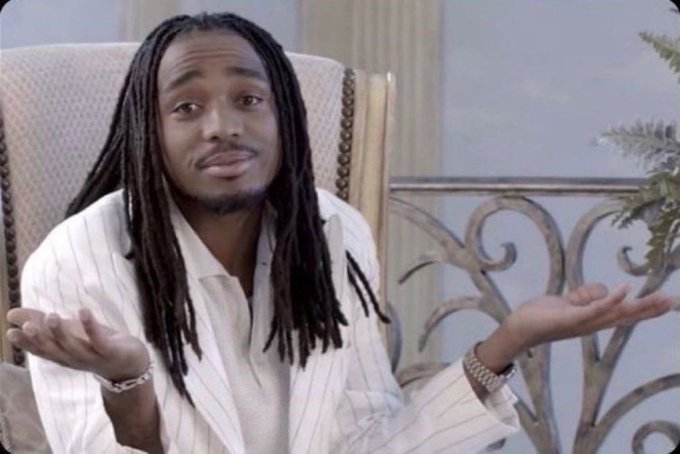 A man with long dreadlocks wearing a white striped shirt sits in an ornate chair and shrugs with his hands raised, palms up, in a gesture of confusion or indifference.
