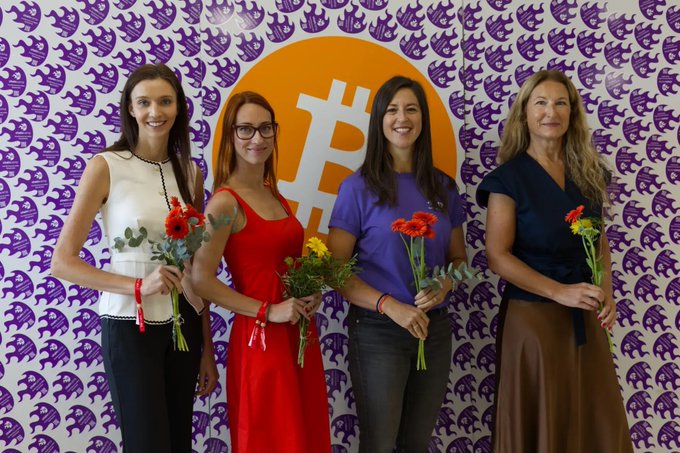 Group photo of four smiling women holding flower bouquets, standing in front of a backdrop patterned with purple Bitcoin logos and a large orange Bitcoin symbol in the center.