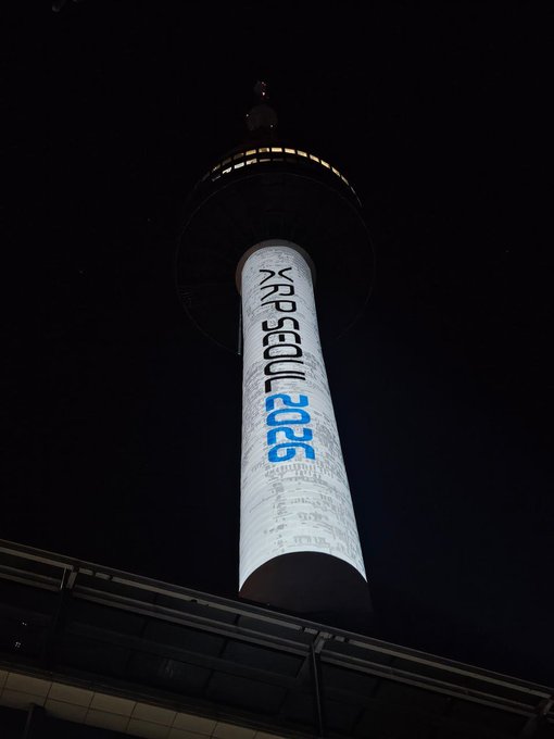 A tall observation tower is lit up at night, displaying a projected logo that reads 'KAP Seoul 2025' along with Korean text down its side.