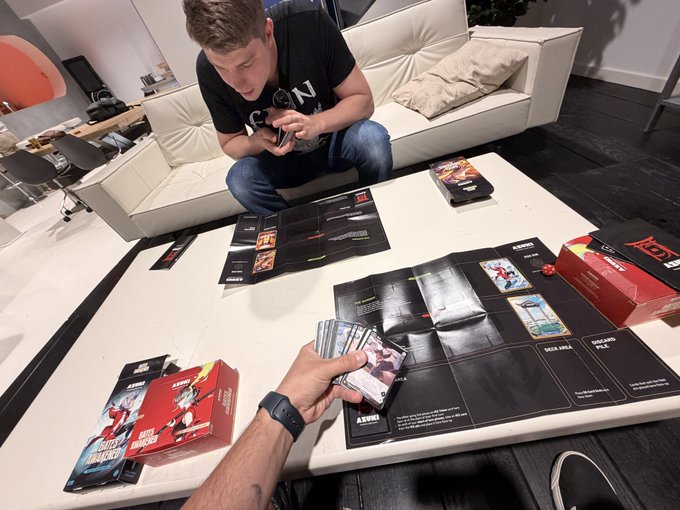 A man in a black t-shirt sits on a white couch holding a camera, photographing someone across a table who is handling cards and boxes related to a tabletop or card game.