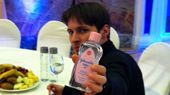 A young man holds up a bottle of Johnson's baby oil toward the camera, with a fruit platter and water bottles visible on the table behind him.