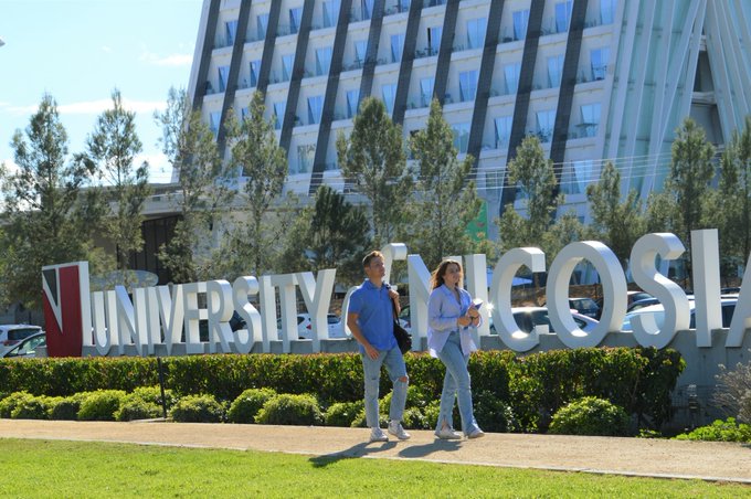 Two people walk past a large 'UNIVERSITY OF NICOSIA' sign on a sunny day, with the modern multi-story building visible in the background.