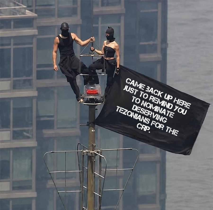 Two people in black balaclavas and black outfits are perched on top of a tall structure holding a large black banner that reads 'CAME BACK HERE JUST TO REMIND YOU TO NOMINATE DESERVING TEZONIANS FOR THE CRP.'